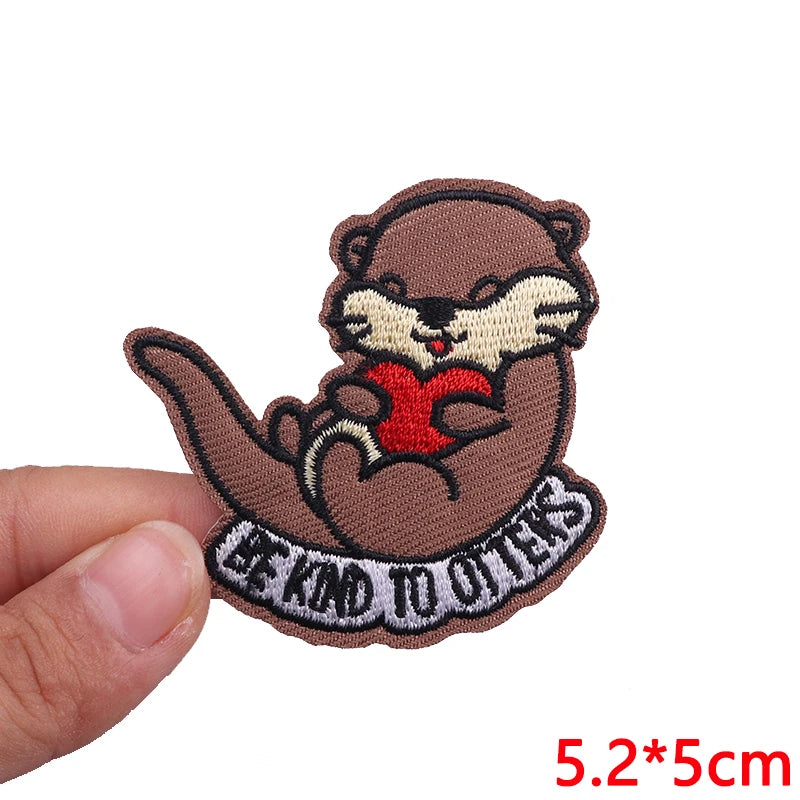 Patch of an otter holding a heart with 'Be Kind to Others' text, held by a hand on a white background.