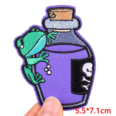 Embroidered patch of a green frog on a purple potion bottle with a skull design, held by a hand.