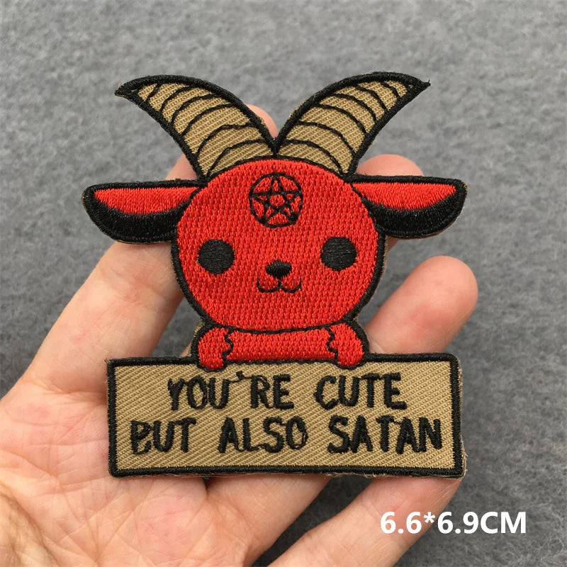 Patch with a cartoonish red creature with horns and a sign saying 'You're cute but also Satan' held in a hand.