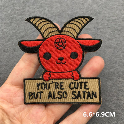 Patch with a cartoonish red creature with horns and a sign saying 'You're cute but also Satan' held in a hand.