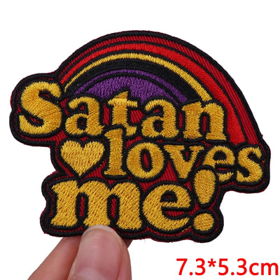 Embroidered patch with 'Satan loves me' text and rainbow design held by a hand.