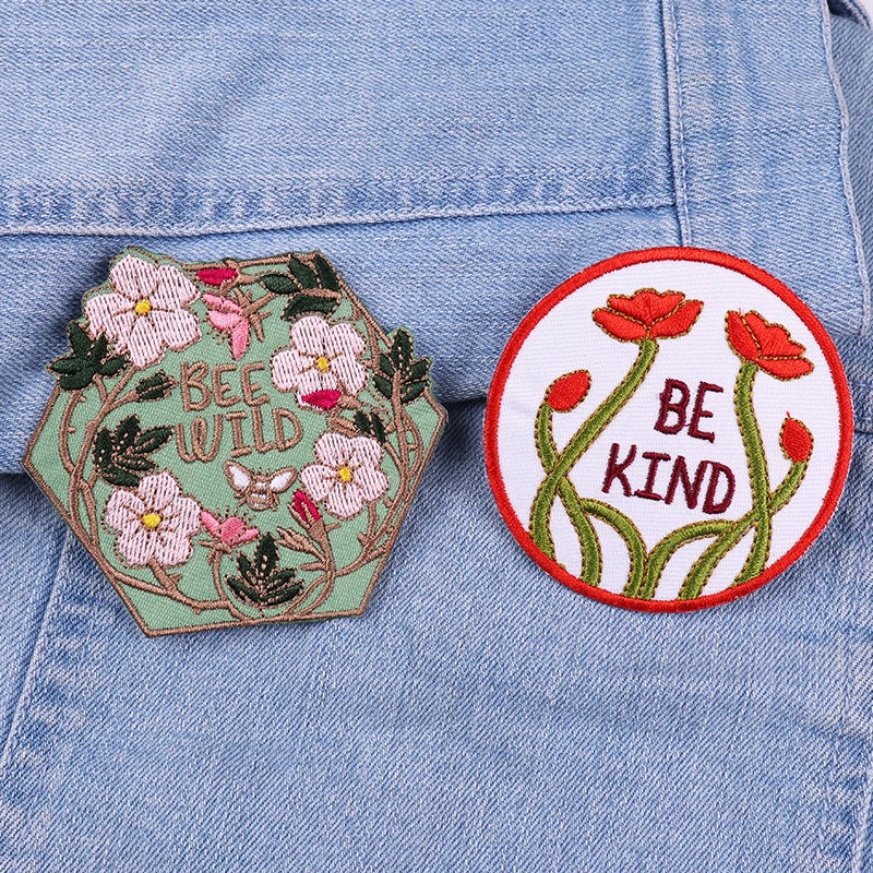 Two embroidered patches on a denim background, one with flowers and 'BEE WILD', the other with red flowers and 'BE KIND'.
