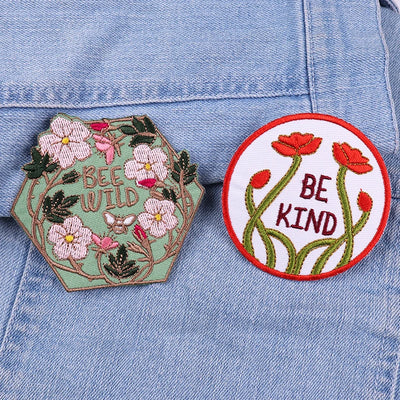 Two embroidered patches on a denim background, one with flowers and 'BEE WILD', the other with red flowers and 'BE KIND'.