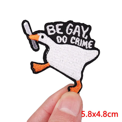 Embroidered patch with a goose holding a bat and text 'BE GAY, DO CAIME' held by a hand.