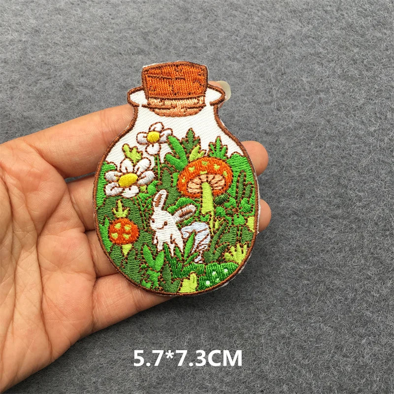 Embroidered patch of a bottle with floral and rabbit design held in hand on gray background