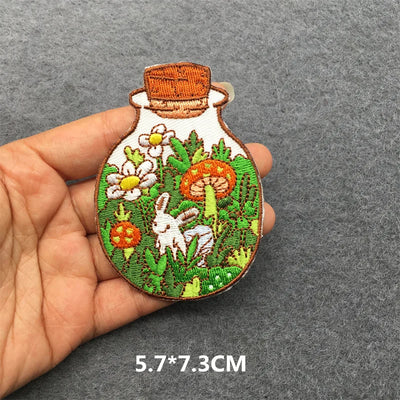 Embroidered patch of a bottle with floral and rabbit design held in hand on gray background