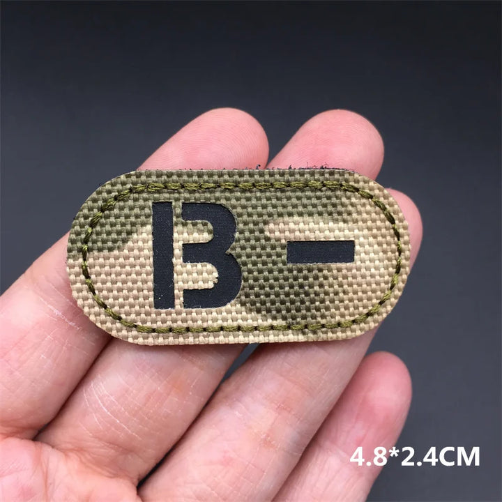 Hand holding an B- multicam IR reflective tactical blood type hook and loop patch approximately 4.8x2.4cm on a dark background