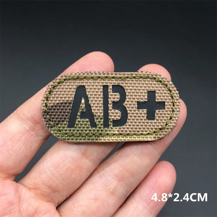 Hand holding an AB+ multicam IR reflective tactical blood type hook and loop patch approximately 4.8x2.4cm on a dark background