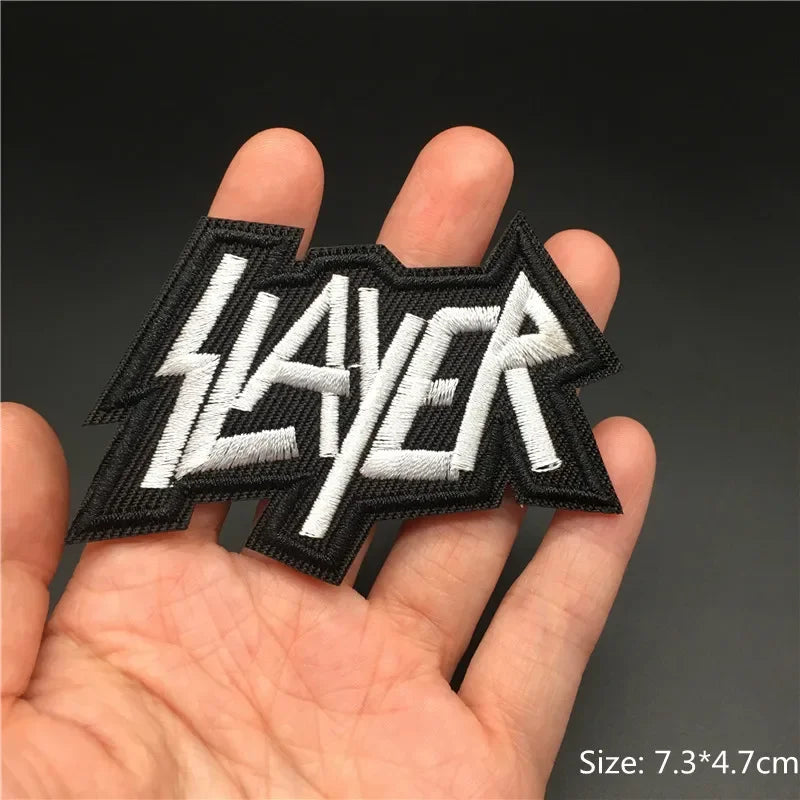 Hand holding a black and white Slayer embroidered iron-on patch 7.3x4.7cm on a dark background
