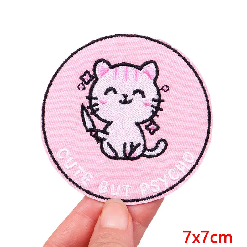 Circular pink patch with a cat illustration and text 'Cute But Psycho' held by a hand on a white background.