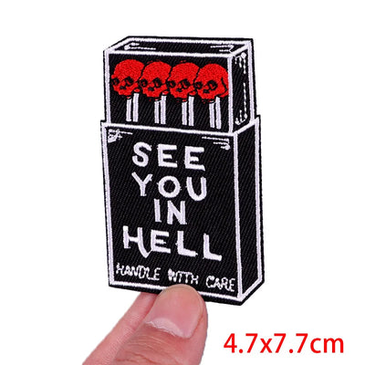 Embroidered patch of a matchbox with skulls and 'See You in Hell' text, held by a hand.