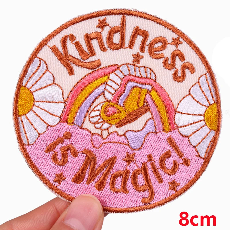 Circular patch with 'Kindness is Magic' text, rainbow, and flowers held by a hand on a white background.