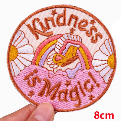 Circular patch with 'Kindness is Magic' text, rainbow, and flowers held by a hand on a white background.