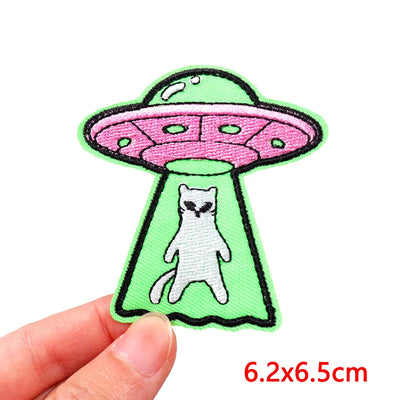 Patch of a cat inside a pink UFO on a green background held by a hand.