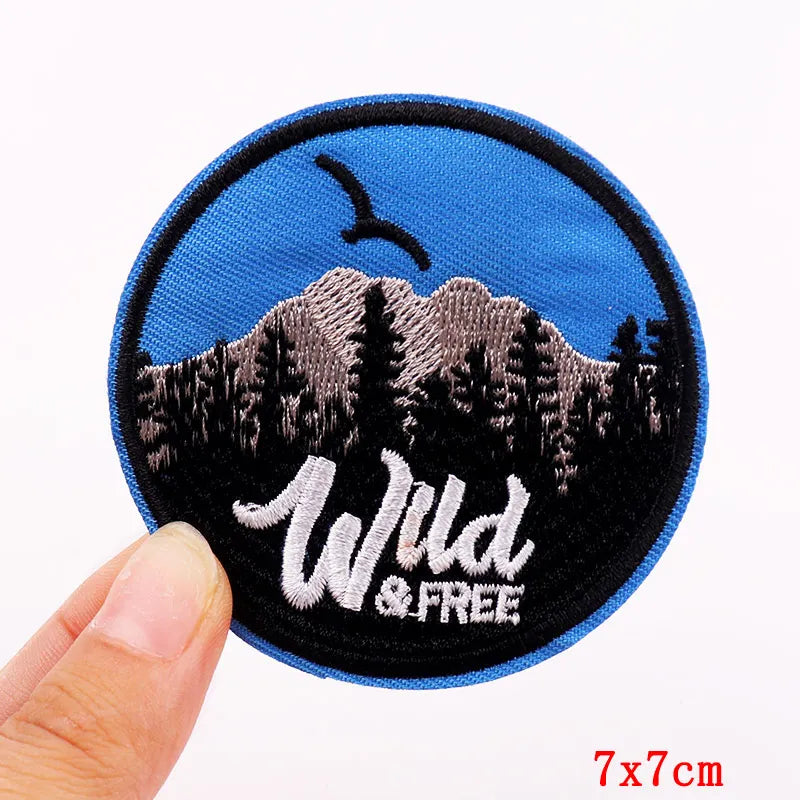 Circular patch with mountain design and 'Wild & Free' text held by a hand on a white background.