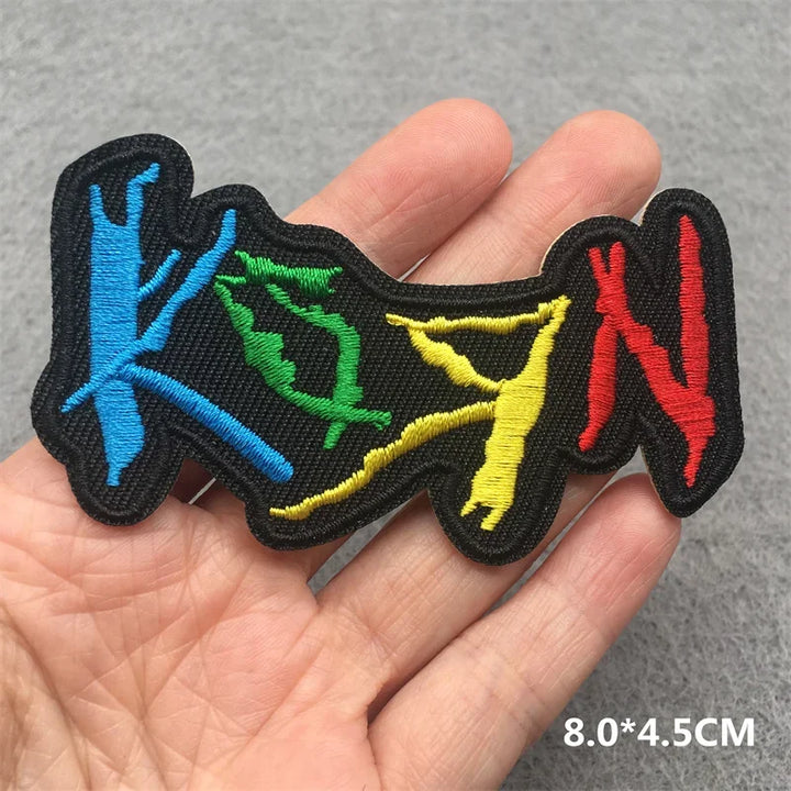 Hand holding a multicolor Korn embroidered iron-on patch 8x4.5cm on a grey background
