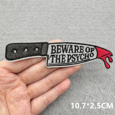 Embroidered patch of a knife with 'Beware of the Psycho' text held in a hand against a gray background.