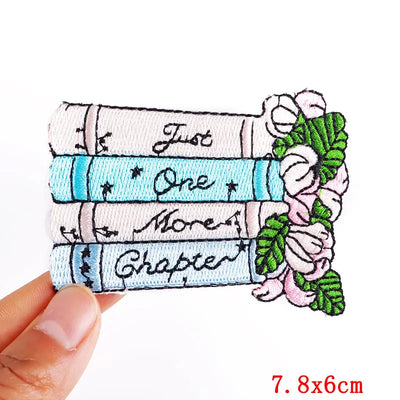 Embroidered patch with books and flowers, held by a hand on a white background.