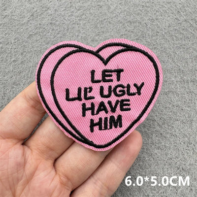 Pink heart-shaped patch with black text held in a hand on a gray background
