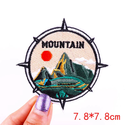 Embroidered patch with mountain design held by a hand, on a white background.