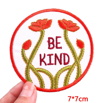 Embroidered patch with 'BE KIND' text and red flowers, held by a hand on a white background.