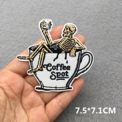Skeleton sitting on a coffee cup patch held in a hand with a gray background