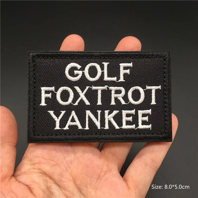 Black patch with 'GOLF FOXTROT YANKEE' text held in a hand against a dark background