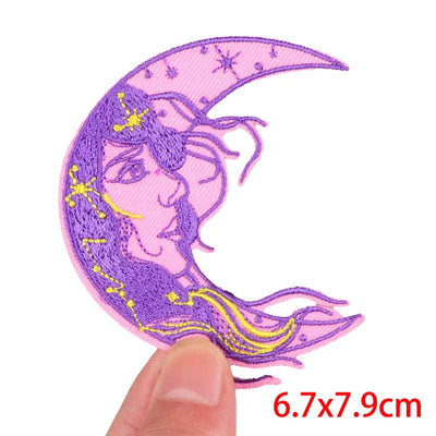 Embroidered patch of a crescent moon with a face, held by a finger, on a white background.