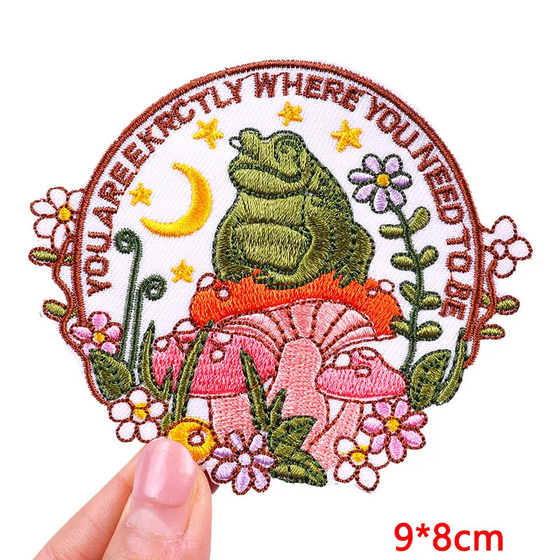 Embroidered patch with a frog, flowers, and text held by a hand on a white background