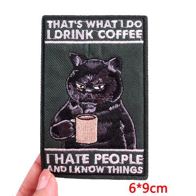 Embroidered patch with a black cat holding a coffee cup and text, held by a hand.