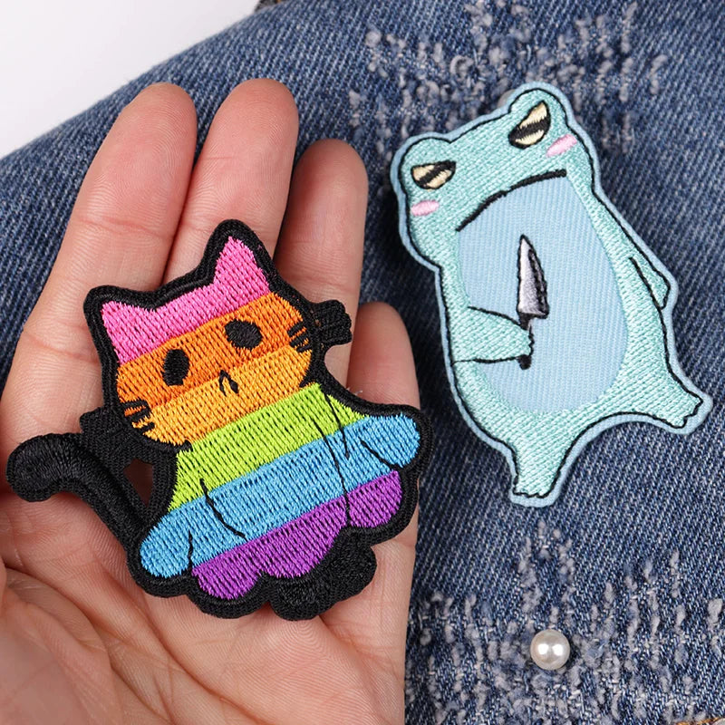 Two colorful patches, one rainbow cat and one blue creature, held against a denim background.