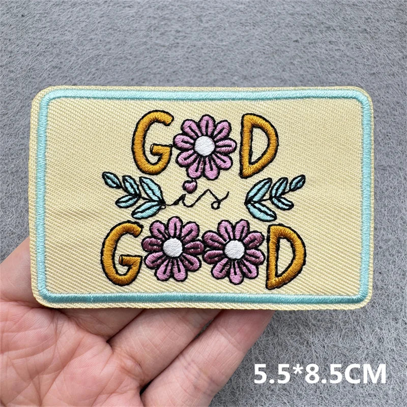 Embroidered patch with 'God is Good' text and flowers held in a hand against a gray background.