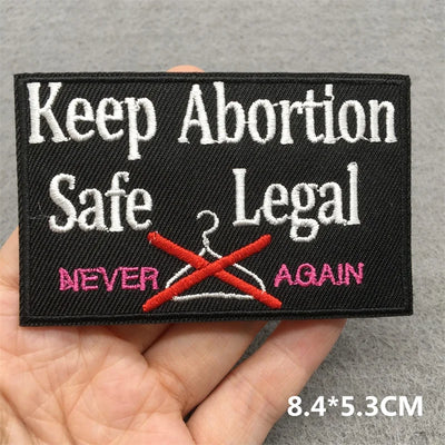 Embroidered patch with text 'Keep Abortion Safe Legal' held in a hand on a gray background.