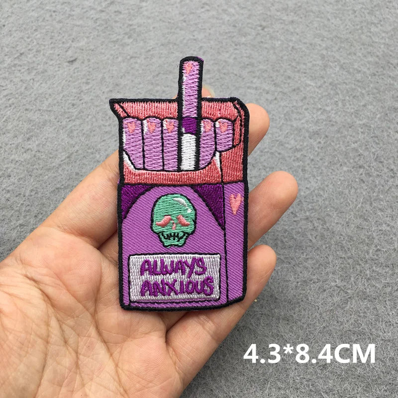 Embroidered patch of a cigarette pack with a hand holding a cigarette, labeled 'Always Anxious', held in a hand against a gray background.