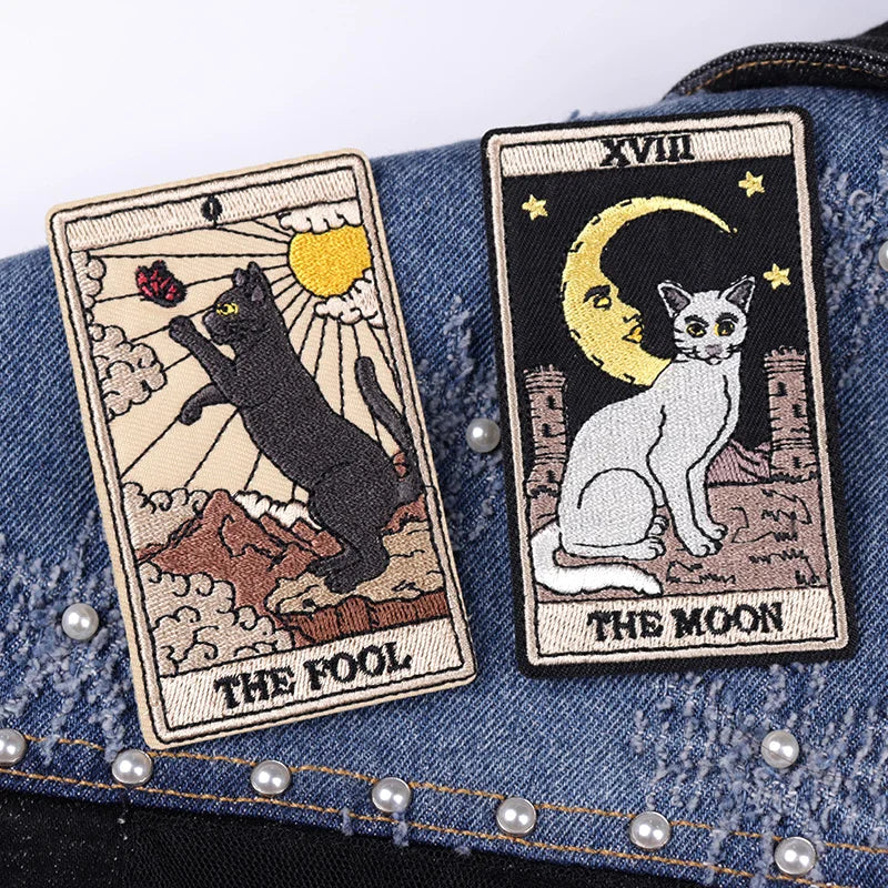 Two tarot card patches on a denim background, featuring 'The Fool' with a black cat and 'The Moon' with a white cat.