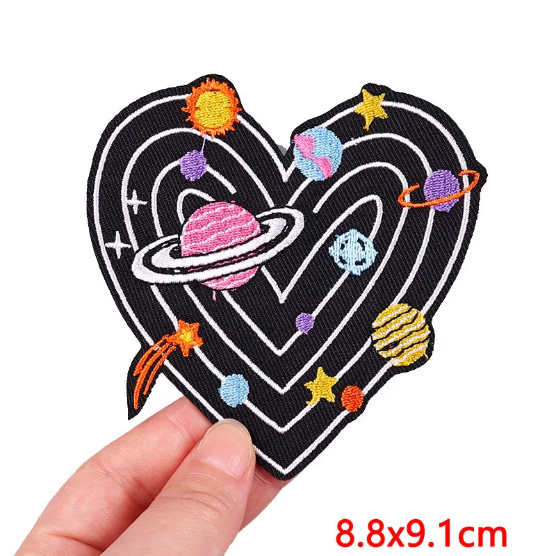 Heart-shaped patch with space-themed design held by a hand, on a white background