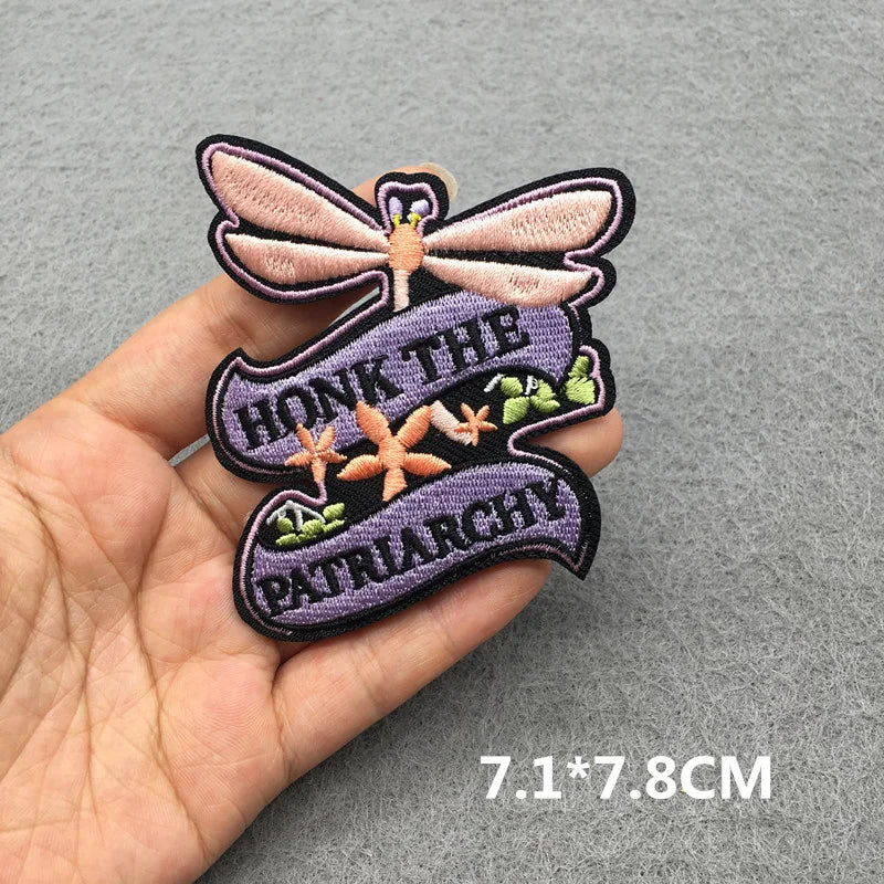 Hand holding a purple pink and black embroidered Honk the Patriarchy dragonfly iron-on patch approximately 7.1x7.8cm on a gray background