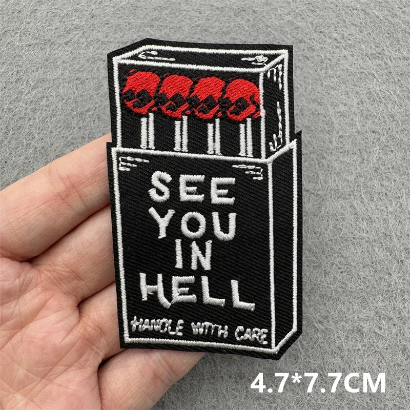 Patch of a matchbox with matches labeled 'See You in Hell' held by a hand on a gray background.