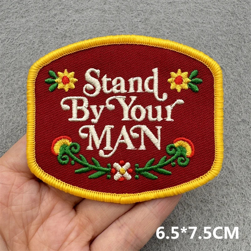 Hand holding a red yellow and green embroidered Stand By Your Man western iron-on patch approximately 6.5x7.5cm on a gray background