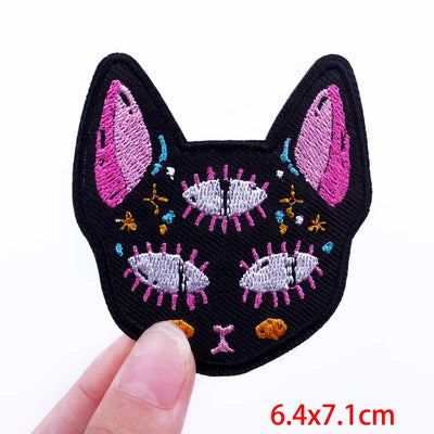 Embroidered patch of a cat's face held by a hand with size measurement '6.4x7.1cm'.