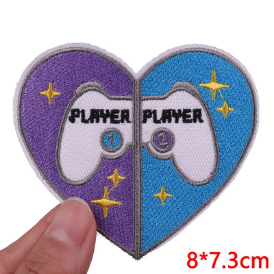 Heart-shaped patch with 'PLAYER' text and gaming controller design, held by a hand.