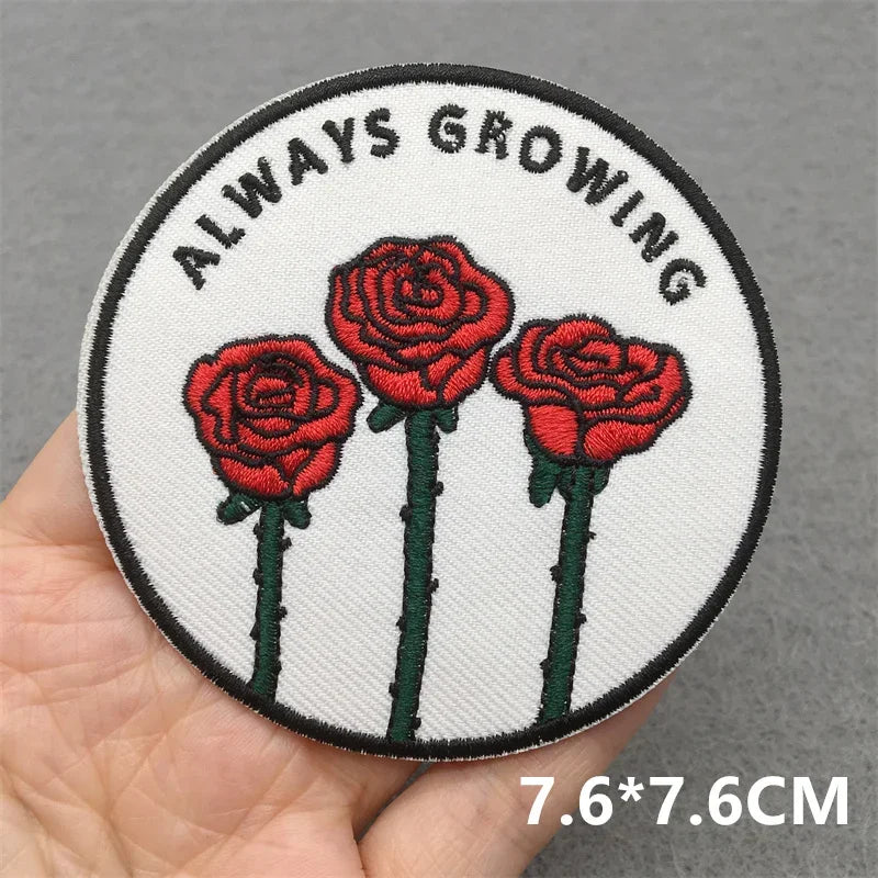 Circular patch with red roses and 'Always Growing' text held by a hand on a gray background.