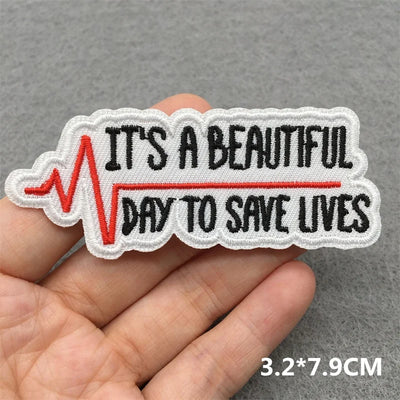 Embroidered patch with 'It's a beautiful day to save lives' text held by a hand on a gray background.