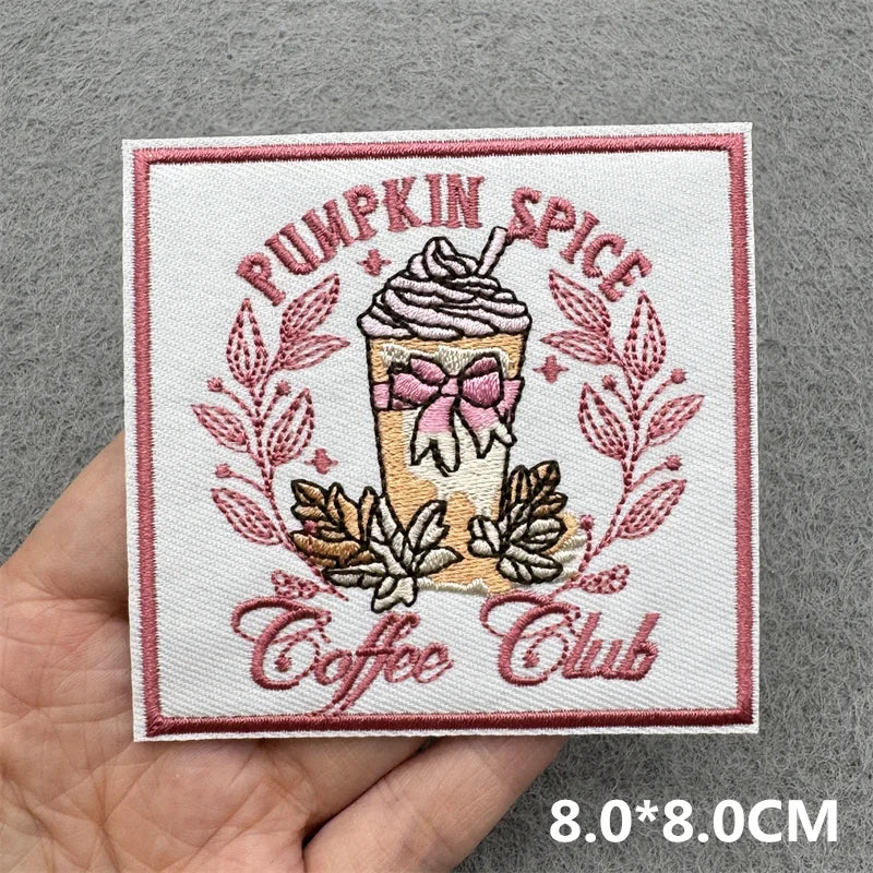 Hand holding a pink cream and brown embroidered Pumpkin Spice Coffee Club iron-on patch with coffee cup and autumn leaves approximately 8x8cm on a gray background