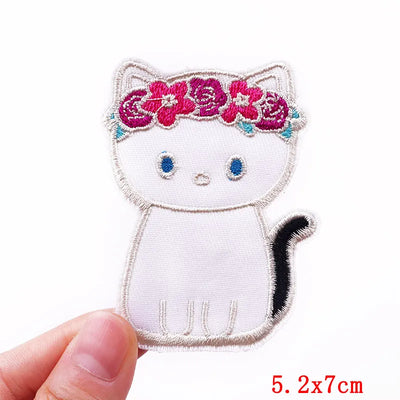 Embroidered patch of a white cat with a pink flower crown held by a hand on a white background.