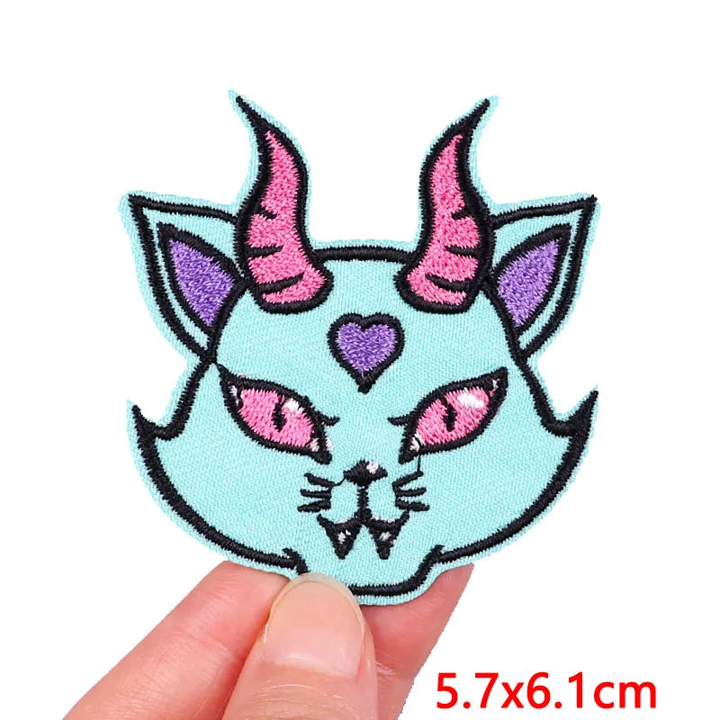 Embroidered patch of a cat face with horns held by a hand, on a white background.