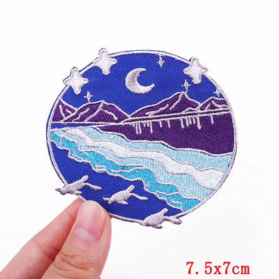 Circular embroidered patch with mountain and landscape design held by a hand, on a white background.