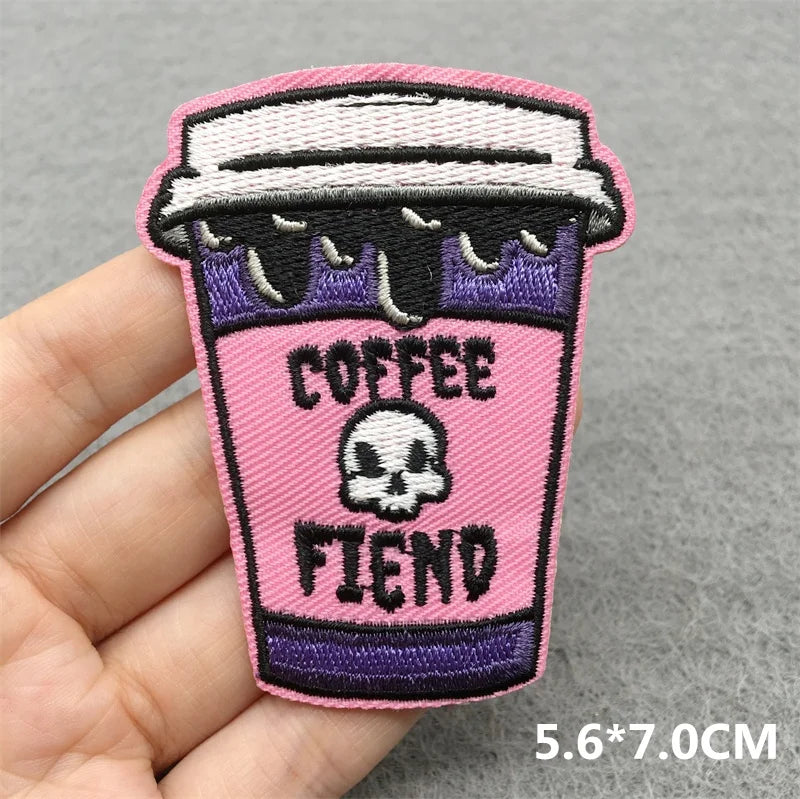 Embroidered patch of a pink coffee cup with a skull design and 'Coffee Fiend' text, held by a hand on a gray background.
