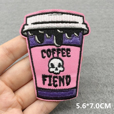 Embroidered patch of a pink coffee cup with a skull design and 'Coffee Fiend' text, held by a hand on a gray background.