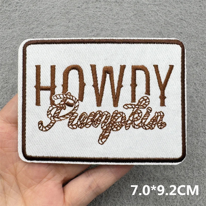 Hand holding a brown and white embroidered Howdy Pumpkin iron-on patch approximately 7x9.2cm on a gray background